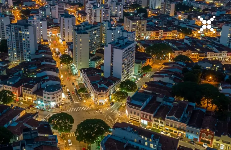 Iluminação Pública Vista aérea de uma cidade brasileira à noite, com ruas bem iluminadas por postes de LED, prédios residenciais e comerciais ao fundo.